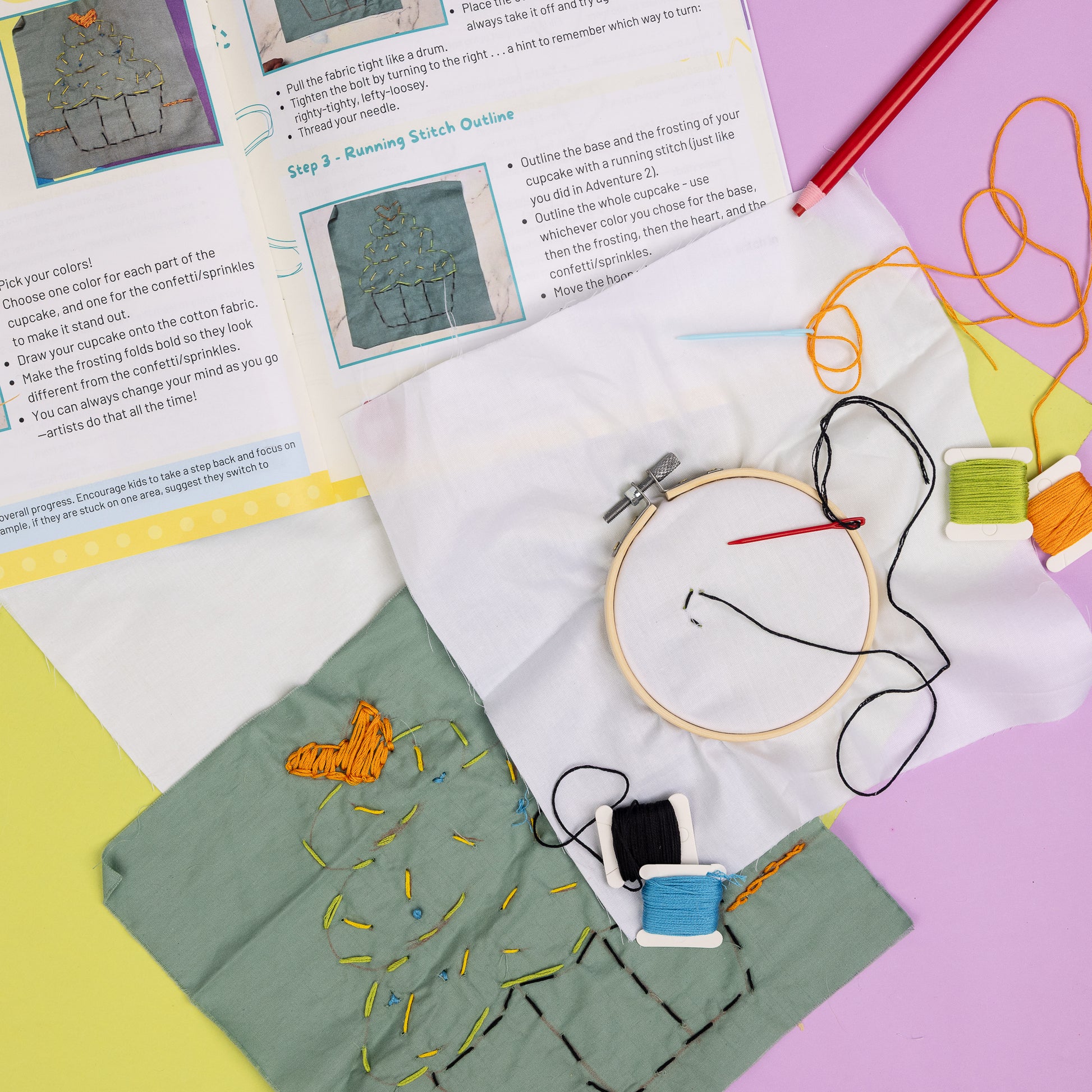 A spread of materials from the Embroidery Art Box including an open-page spread from the instructions, embroidery hoop with cotton fabric, needle, and thread, additional thread and needles, fabric pencil, and final project of an embroidered cupcake on cotton fabric.
